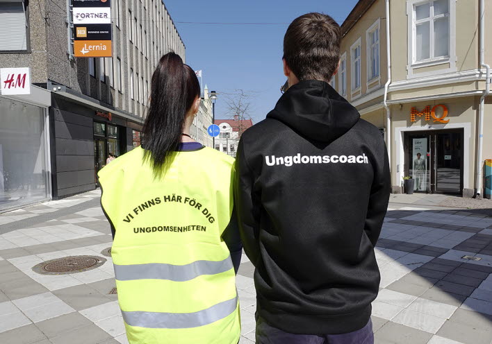 Två personer vända med ryggen mot kameran. en person bär skyddsväst med texten Vi finns här för dig, ungdomsenheten. På den andra personens hoodie står det Ungdomscoach.