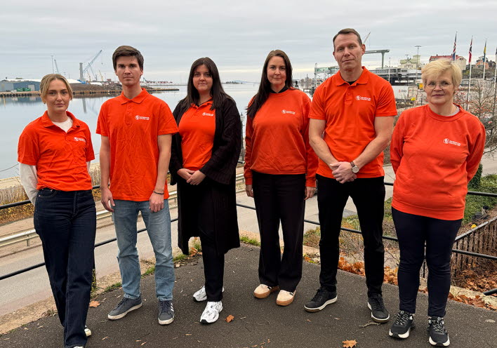 Sex medarbetare i orangea tröjor står på en balkong med hamnen i bakgrunden.