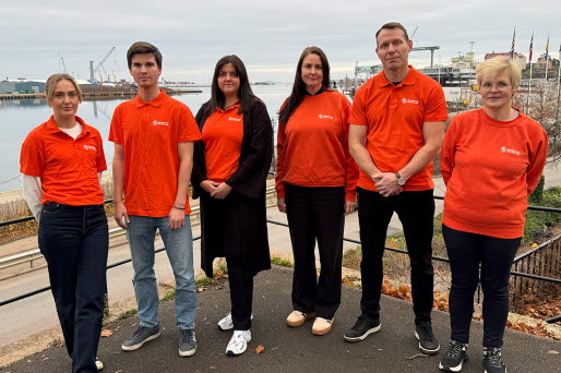 Sex medarbetare i orangea tröjor står på en balkong med hamnen i bakgrunden.