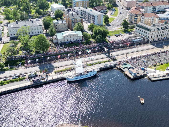 Vy över hamnen med segelbåt och publik längs kajkanten. 