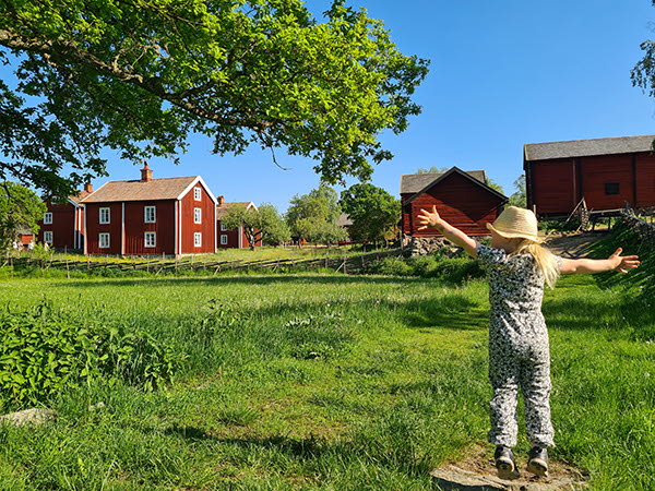 Ett barn med armarna utsträckta i ett somrigt landskap med gräs och röda hus.